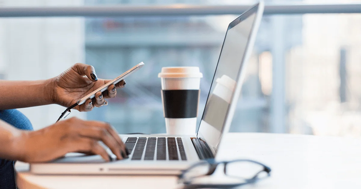 image of an open laptop, a woman's hand on the keyboard, her other hand holding a smartphone. It is daylight, there are a pair of glasses beside the computer in the foreground and a takeout coffee in the background. The image is by Christina @ wocintech on Unsplash.