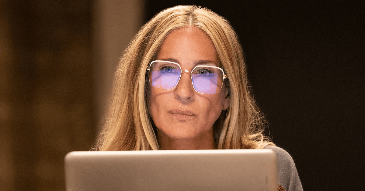 Craig Blankenhorn/HBO image of Sarah Jessica Parker in the Sex and the City Reboot And Just Like That... Carrie Bradshaw, with straight hair and glasses, sits behind an open Apple MacBook, the screen reflected in her lenses. The background is dark.