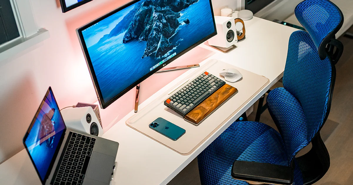 Overhead picture of a home workstation including wireless mouse and keyboard, laptop on a stand, external speakers, widescreen monitor, and wall mounted television, with a fancy blue office chair.